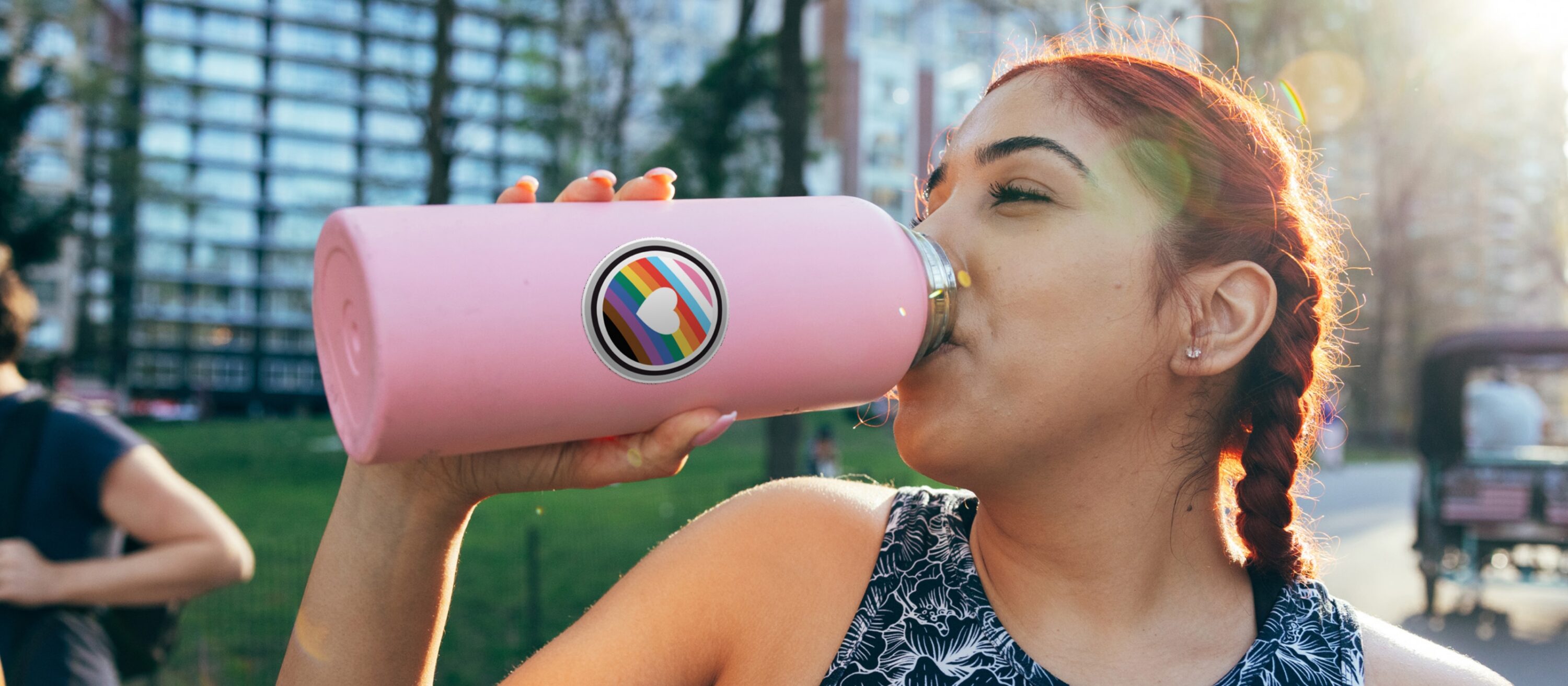 Person drinking from a pink reusable water bottle with a rainbow heart sticker outdoors.
