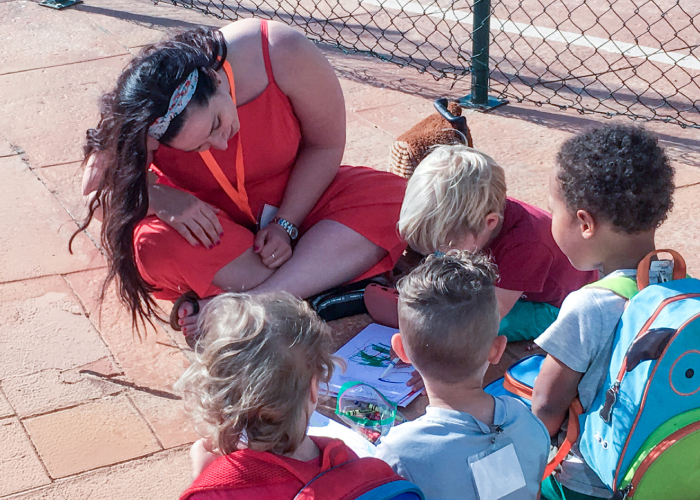 img-sn-community2-700-1 Community volunteer sitting with young children outdoors while guiding them through a drawing activity.