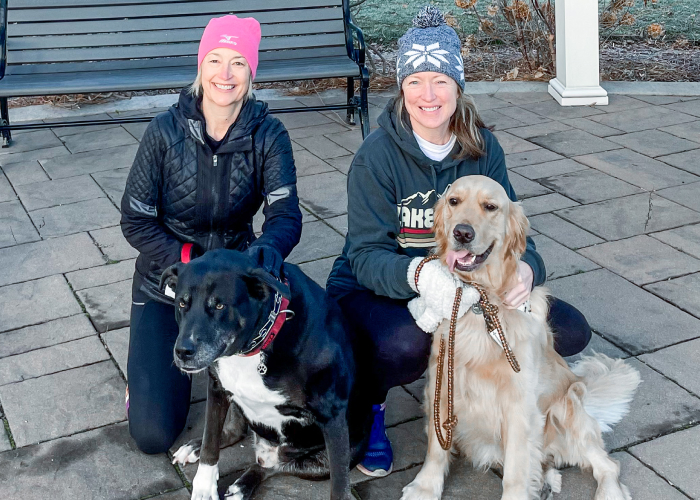 img-sn-community5-700-1 Two women kneeling outdoors with their dogs, smiling together in a park setting.